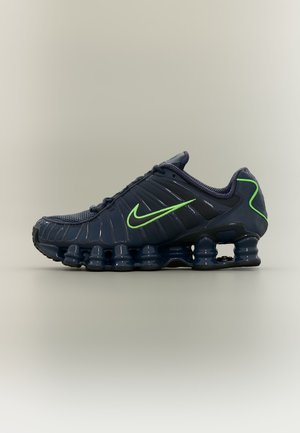 Sötétkék Nike futócipő neon zöld logóval és rugószerű lengéscsillapítókkal a talpon, oldalnézetben.