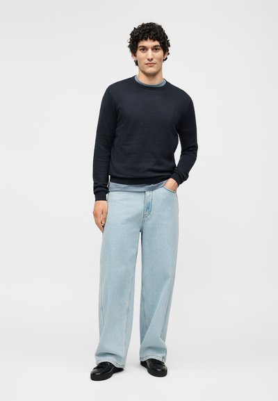 Νέος άνδρας με σγουρά μαλλιά, φοράει μαύρο πουλόβερ, ανοιχτό μπλε τζιν με φαρδιά μπατζάκια και μαύρα παπούτσια, στέκεται με το ένα χέρι στην τσέπη.