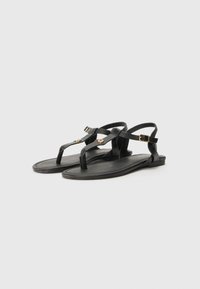 Sandalias planas negras con un diseño de tira en T, hechas de cuero suave. Presentan correas en el tobillo con hebillas y sutiles acentos dorados en las correas.
