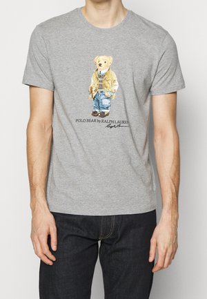 Mężczyzna w szarej koszulce z nadrukiem niedźwiedzia ubranego w warstwowe, casualowe ubrania, pod którym znajduje się napis „POLO BEAR by RALPH LAUREN”.
