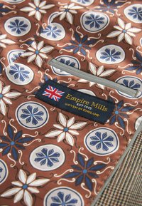 Brun mønstret tekstil med blomster motiver i hvid og blå. Etiket læser "Empire Mills, EST 1915, VEVET I ENGLAND" med et Union Jack.