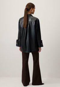 Sort faux læderskjorte med krave, lange ærmer med splitmanchetter. Modellen er iført brune flared bukser og står med ryggen til kameraet.