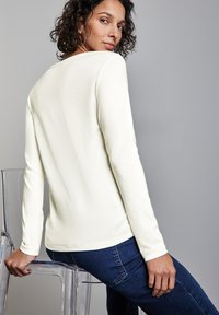 Långärmad, krämfärgad topp med mjuk textur och avslappnad passform. Kombinerad med mörkblå denimjeans. Klar plaststol i bakgrunden.