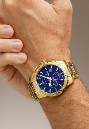 Gold metal wristwatch with blue dial, chronograph subdials, date display, and rotating bezel worn on left wrist.