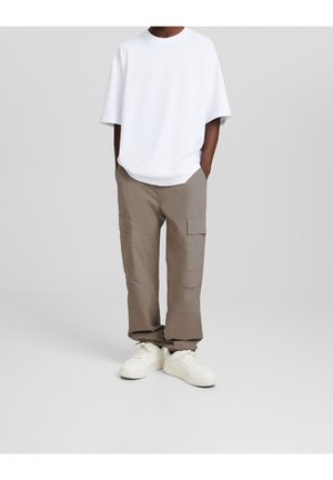 Man som bär en vit oversized t-shirt, bruna cargobyxor med fickor och vita sneakers, stående med händerna i fickorna mot en enfärgad bakgrund.