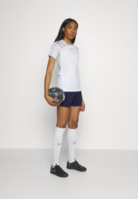 Nike športový set pozostávajúci z bieleho trička s krátkym rukávom a šedými detailmi, spolu s ladiacimi tmavomodrými šortkami. Čierne futbalové topánky a biele ponožky po kolená.