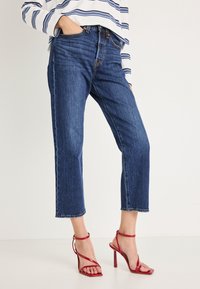 Τζιν παντελόνια σε σκούρο μπλε, με ίσια γραμμή και ακατέργαστο τελείωμα· συνδυασμένα με κόκκινες ψηλοτάκουνες σανδάλες με λουράκια, που αναδεικνύουν το ανοιχτό σχέδιο στο πέλμα.