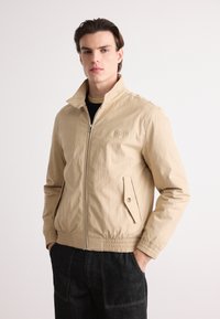 Chaqueta ligera beige con cuello alto, cierre de cremallera, bolsillo lateral y puños elásticos. Textura suave y ajuste casual.