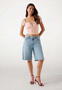 Top corto rosa chiaro con maniche a sbuffo e dettagli in pizzo; bermuda in denim blu con orlo sfrangiato e vestibilità rilassata; scarpe con tacchi a strappo color bordeaux.