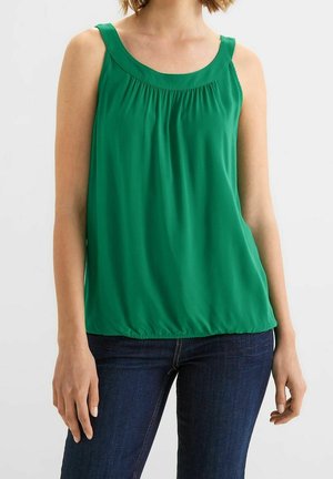 Vrouw draagt een mouwloze groene blouse met geplooide halslijn en donkerblauwe spijkerbroek, staand tegen een effen lichte achtergrond.