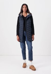 Gevoerde marineblauwe vest met capuchon, met een contrasterende binnenvoering, gedragen over een zwarte gebreide truien, gecombineerd met lichtblauwe spijkerbroek en beige schoenen.