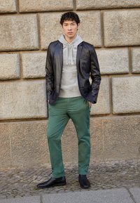 Černá kožená blazer přes šedou mikinu, doplněné zelenými kalhotami a černými botami. Hladké textury, strukturovaný design a kontrastní barvy.