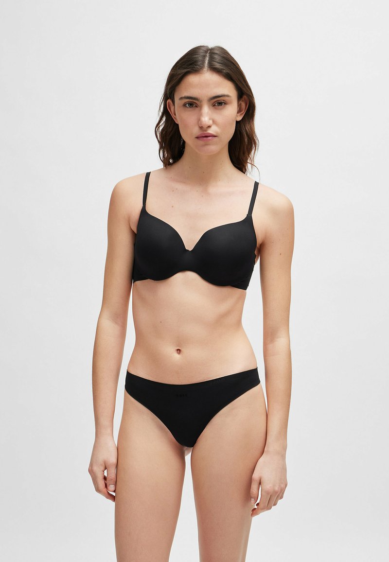 Frau mit langen braunen Haaren, die ein passendes Set aus schwarzen BH und Unterwäsche trägt, steht vor einem schlichten hellgrauen Hintergrund.