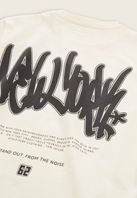 Bijela pamučna majica s odvažnim crnim grafitnim tekstom. Tekst uključuje fraze i brojeve, s glatkom teksturom tkanine.
