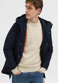 Chaqueta acolchada azul marino con capucha, con un forro naranja, combinada con un suéter de punto trenzado en color crema y jeans azules.