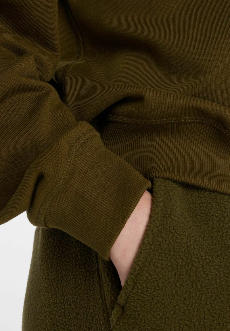 Sweat à capuche en polaire vert olive avec poignets côtelés. Gros plan sur une main dans la poche montrant la texture douce et les éléments de tissu contrastants.