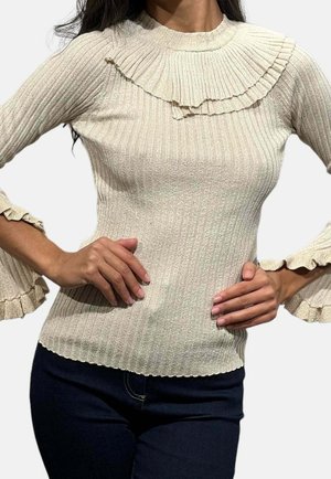 Žena v béžovém žebrovaném svetru s volánovým límcem a rozšířenými manžetami, v kombinaci s tmavými kalhotami, ruce opřené o boky.