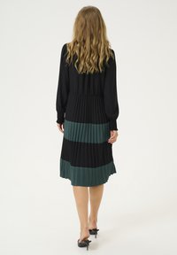 Melna un zaļa plāksnīte midi kleita ar garām piedurknēm, savilktu vidukli un plūstošu hemu; satur kontrastējošus auduma lentes un teksturētu dizainu.