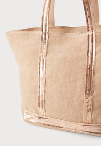 Šviesiai rudos drobės rankinė su blizgiomis rožinės aukso pailgomis diržais, turinti erdvų atidarymą ir lygią bazę stabilumui.