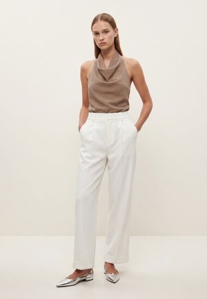 Žena u bezrukom topu boje taupe s ovratnikom, bijelim širokim hlačama s visokim strukom i srebrnim špicnatim balerinkama, stoji s rukama u džepovima.