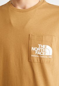 Ruda trumpa rankovė t-shirt marškiniai, pagaminti iš medvilnės, su baltu logotipo spaudiniu kairėje krūtinės pusėje ir apvaliu kaklu.