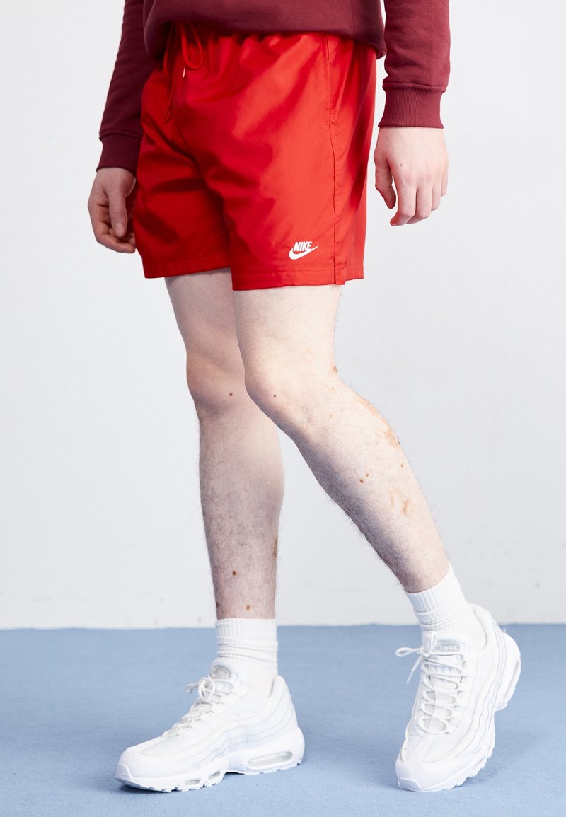 Personne portant un short Nike rouge, des chaussettes blanches et des chaussures de sport blanches debout sur un sol bleu devant un fond uni.