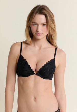 Frau mit langen blonden Haaren, die einen schwarzen Spitzen-BH trägt, steht vor einem beigen Hintergrund und blickt mit neutralem Ausdruck nach vorne.