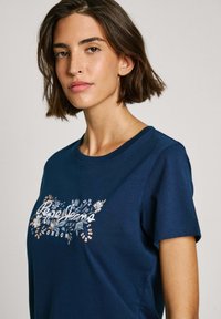T-shirt à manches courtes bleu marine en coton, avec un imprimé floral et le texte "Pepe Jeans London" en couleurs contrastées.