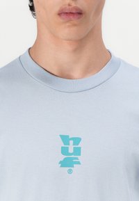 Svetlo modré tričko s krátkym rukávom, s výrazným tyrkysovým logom na hrudi. Bavlnený materiál s rebrovaným okrúhlym výstrihom.