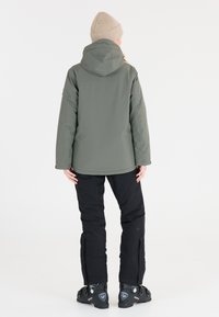 Grüne wasserdichte Jacke mit Kapuze, kombiniert mit schwarzen Schneehosen und dunklen Stiefeln. Die Jacke hat eine glatte Textur und minimale Designdetails.
