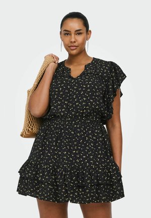 Kobieta w czarnej kwiecistej sukience z falbankami trzyma plecioną, beżową torbę na ramieniu na jednolitym tle.