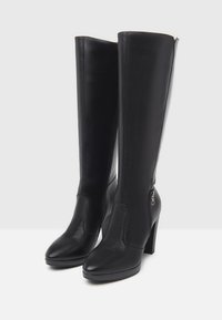 Bottes en cuir noires montant jusqu'au genou, avec un bout pointu, un talon haut et une fermeture éclair latérale. Présentent une texture lisse et des détails de couture contrastés.