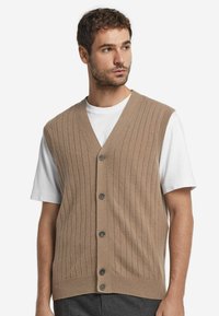 Hellbrauner, gerippter Strickweste mit V-Ausschnitt, ausgestattet mit fünf Knopfverschlüssen, über einem weißen T-Shirt getragen; strukturierter Stoff und lässiges Design.