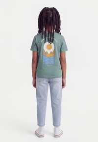 Παιδί με μακριά σγουρά μαλλιά στέκεται με την πλάτη, φοράει πράσινο μπλουζάκι με γραφικό ήλιου, βουνών και νερού, ανοιχτόχρωμο τζιν και λευκά αθλητικά παπούτσια.