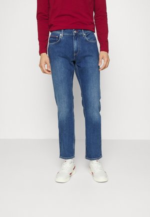 Man wearing blue denim jeans, white sneakers, and a long-sleeve red shirt, standing against a plain light background.