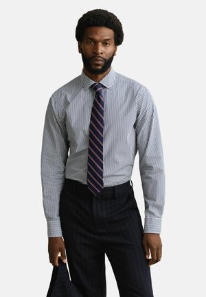 Mann mit Bart, der ein gestreiftes Hemd, eine marineblaue gestreifte Krawatte und eine dunkle Nadelstreifenhose trägt und die passende Jacke in der Hand hält.