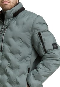 Zimowa kurtka ocieplana w kolorze zielonym, z wysokim kołnierzem, zamykanymi na zamek kieszeniami i teksturowaną powierzchnią. Posiada czarne akcenty oraz naszywkę z logo na rękawie.