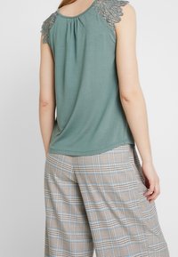 Grön ärmlös topp med spetsdetaljer på axlarna, parat med beigefärgade rutiga byxor med vida ben som har blå accentfärger och en avslappnad passform.