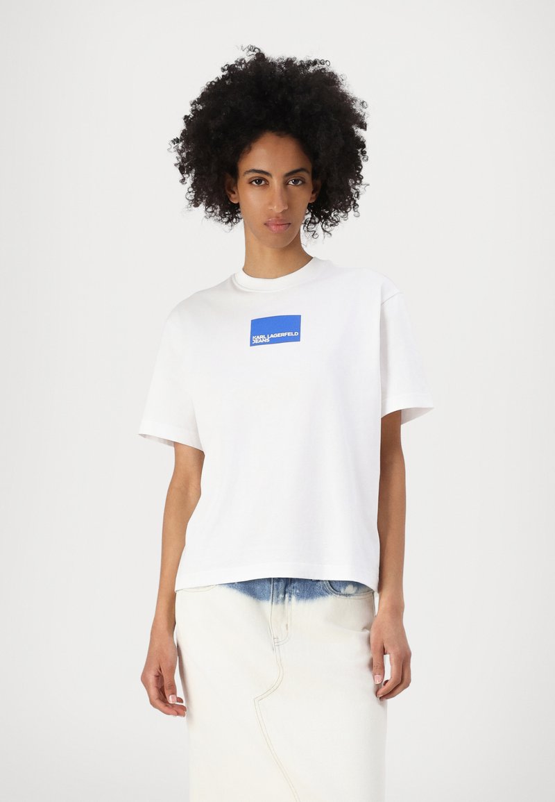 Tricou alb din bumbac cu mâneci scurte, având o etichetă logo rectangulară albastră pe piept. Asortat cu o fustă din denim deschis.