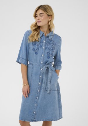 Femme portant une robe en jean bleu longueur genou avec broderie florale, boutonnée à l'avant, manches courtes, ceinture à la taille et poches, debout les mains détendues.