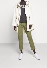 Bílá nepromokavá bunda se stahovacími šňůrkami, šedé tričko s velkým nápisem, olivově zelené krátk é kalhoty a bílé tenisky. Jemný, ležérní vzhled.