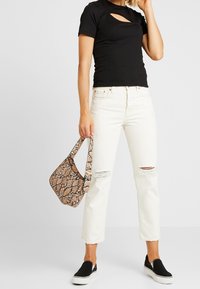 Kvinna i svart cutout-topp, vit ripped jeans, svarta slip-on skor och håller en liten handväska i beige med snakeskin-mönster.