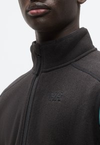 Γιλέκο από φλις σε σκουρό γκρι χρώμα, με ψηλό γιακά, φερμουάρ κατά μήκος ολόκληρης της μπροστινής πλευράς και λογότυπο στο στήθος. Το υλικό φαίνεται μαλακό με υφή.