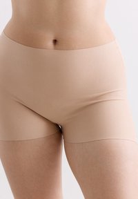 Nahaufnahme des Mittelsections einer Person in nahtlosen beigefarbenen Shorts vor einem neutralen Hintergrund.