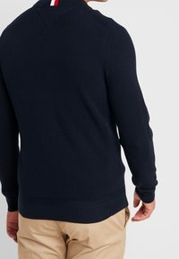 Námořnický modrý pletený pulovr s texturovaným vzorem, se správou červeného a bílého pruhu na límci a žebrovanými manžetami. Klasický střih.