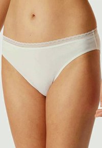 Donna che indossa biancheria intima di cotone bianco con pizzo in vita, mostrando il torso e la parte superiore delle cosce su uno sfondo semplice.