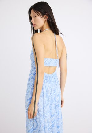 Młoda kobieta z długimi ciemnymi włosami, ubrana w niebieską, odkrytą z tyłu sukienkę na halter, stojąca bokiem na jednolitym białym tle.