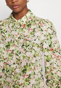 Květinová halenka s krémovým pozadím, růžovými a zelenými abstraktními vzory, špičatým límcem a zapínáním na knoflíky vpředu.