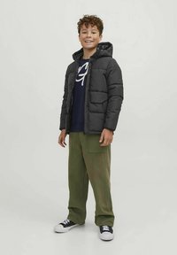 Melna pūkaina jaka ar kapuci pār tumši zilu kreklu ar baltu uzrakstu, kopā ar olīvdzeltenām biksēm un melniem sporta apaviem.