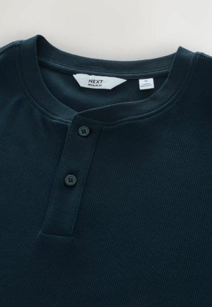 Tumši zili riboti henley krekls ar apaļu kakla izgriezumu, divām melnām pogām un etiķeti, kas norāda vidējo izmēru no NEXT.